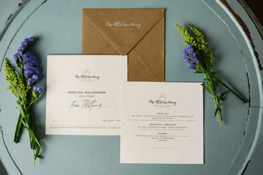 Hochzeitseinladungssuite mit zwei Karten und einem braunen Umschlag auf einem hellblauen runden Tisch mit floralen Akzenten. Der deutsche Text zeigt die Kompetenz von Grafik Design München mit eleganter schwarzer und goldener Schrift für ein raffiniertes visuelles Branding.