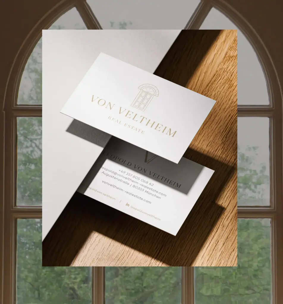Zwei Visitenkarten für Von Veltheim Real Estate, gestaltet mit Grafik Design München, werden auf einer Holzfläche im Sonnenlicht präsentiert. Die obere Karte zeigt das Firmenlogo und den Namen, die untere Karte die Kontaktdaten von Leopold Von Veltheim.