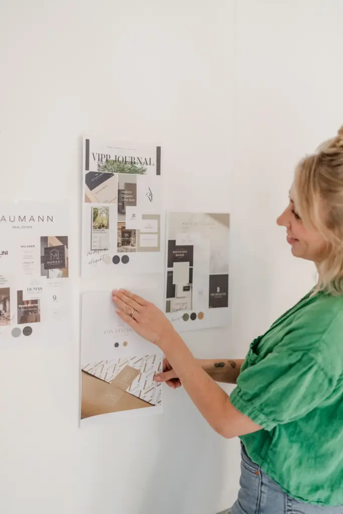 Grafikdesignerin Carina Zwerenz in einem grünen Hemd lächelt, während sie Moodboards und Designmaterialien mit visuellen Branding-Elementen an eine weiße Wand pinnt. Die Tafeln zeigen Bilder, Farbmuster und Texte für Corporate Design München Projekte.