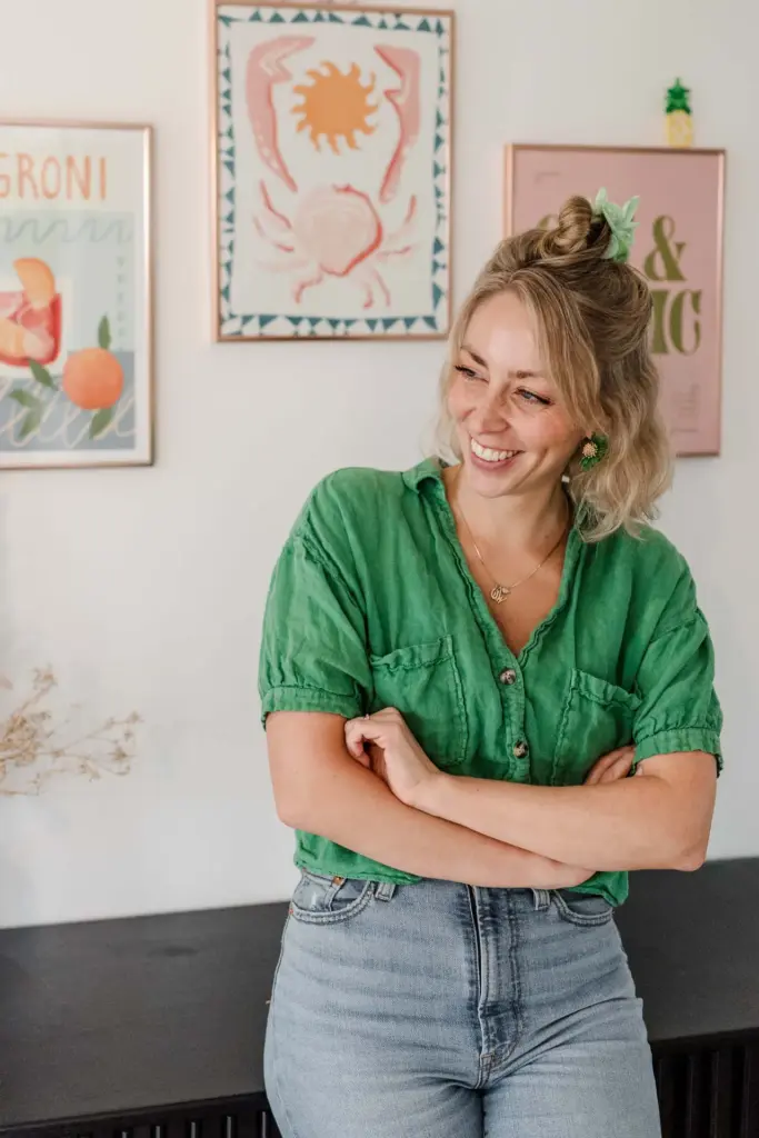 Eine Frau mit blonden, hochgesteckten Haaren, Grafikdesignerin Carina Zwerenz, steht lächelnd und mit verschränkten Armen da, sie trägt ein grünes Hemd und hellblaue Jeans. An der Wand hinter ihr hängt ein buntes gerahmtes Kunstwerk, das visuelles Branding und Grafik Design München widerspiegelt.