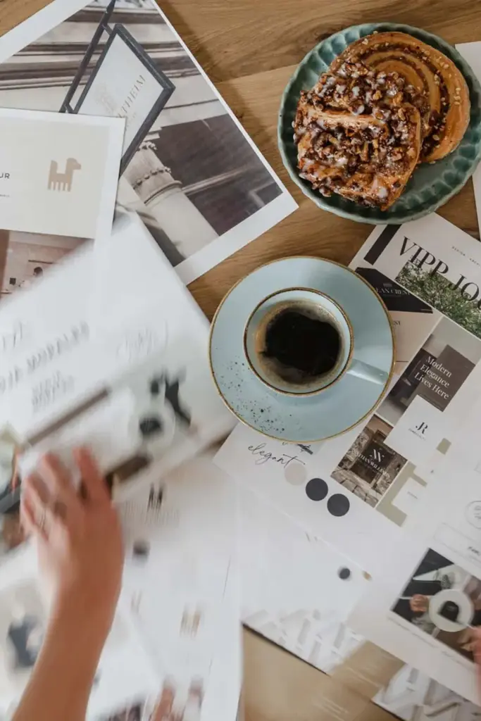 Ein Tisch mit verstreuten Papieren und Designdrucken deutet auf logodesign München hin, während eine blaue Tasse mit schwarzem Kaffee und Gebäck die Szene vervollständigt. Eine Person streckt die Hand aus, vertieft in visuelle Branding-Ideen.