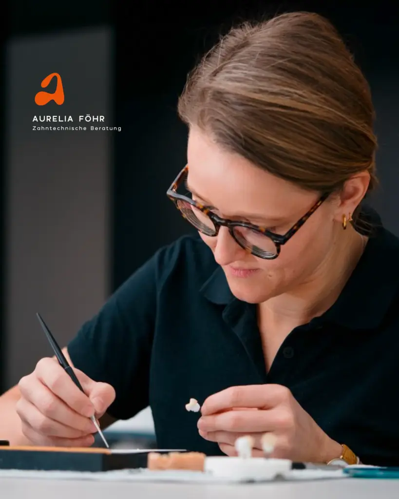 Eine Frau mit Brille und schwarzem Hemd arbeitet sorgfältig an einer Zahnform, wobei sie ein Werkzeug und ein Zahnersatzstück hält. Auf der linken Seite des Bildes erscheinen ein visuelles Branding und das Logo von Aurelia Föhr Zahntechnische Beratung.